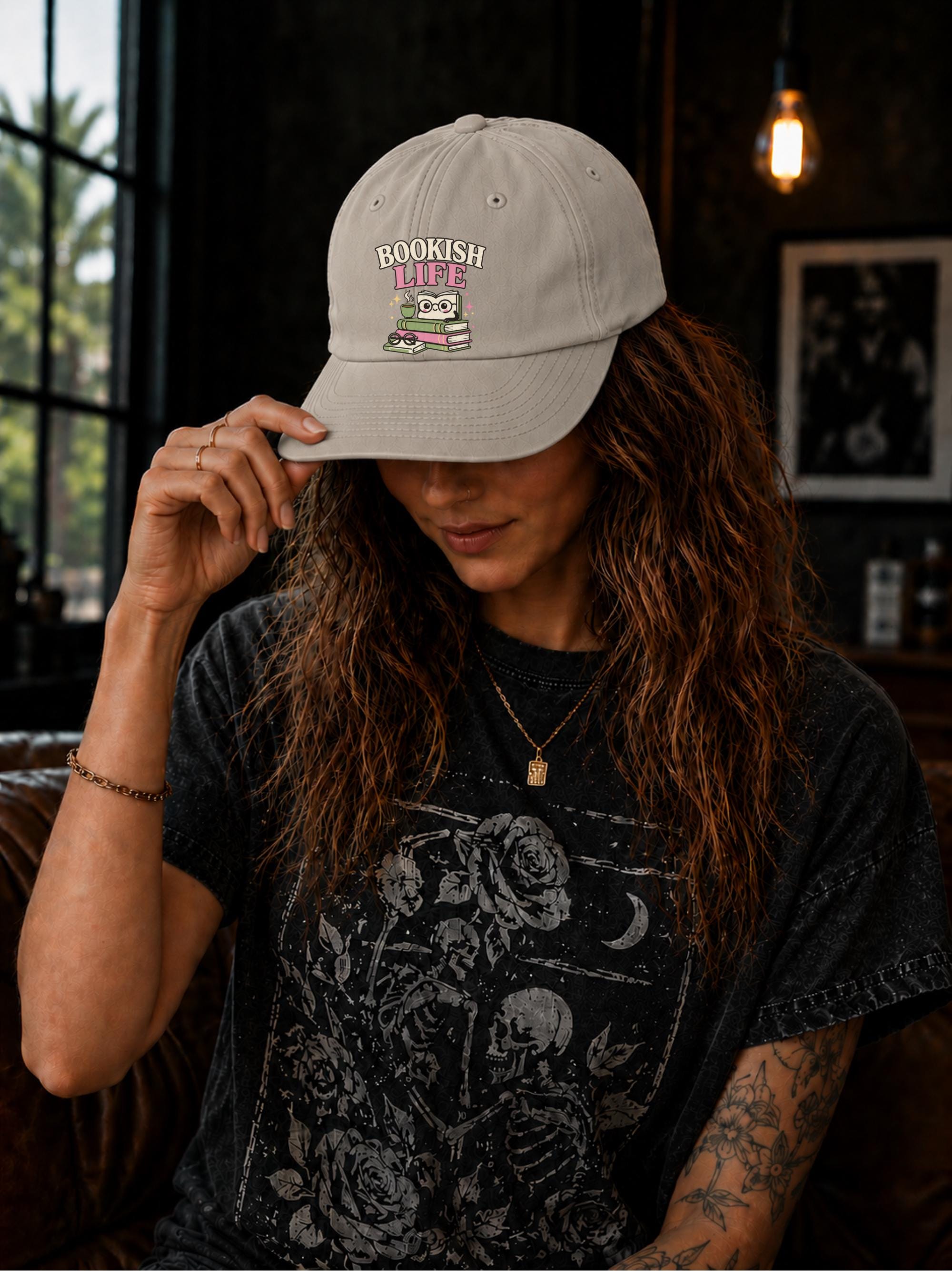 Woman wearing a stone classic dad cap featuring a colorful Bookish Life graphic with stacked books, glasses, and a playful reading-themed illustration.