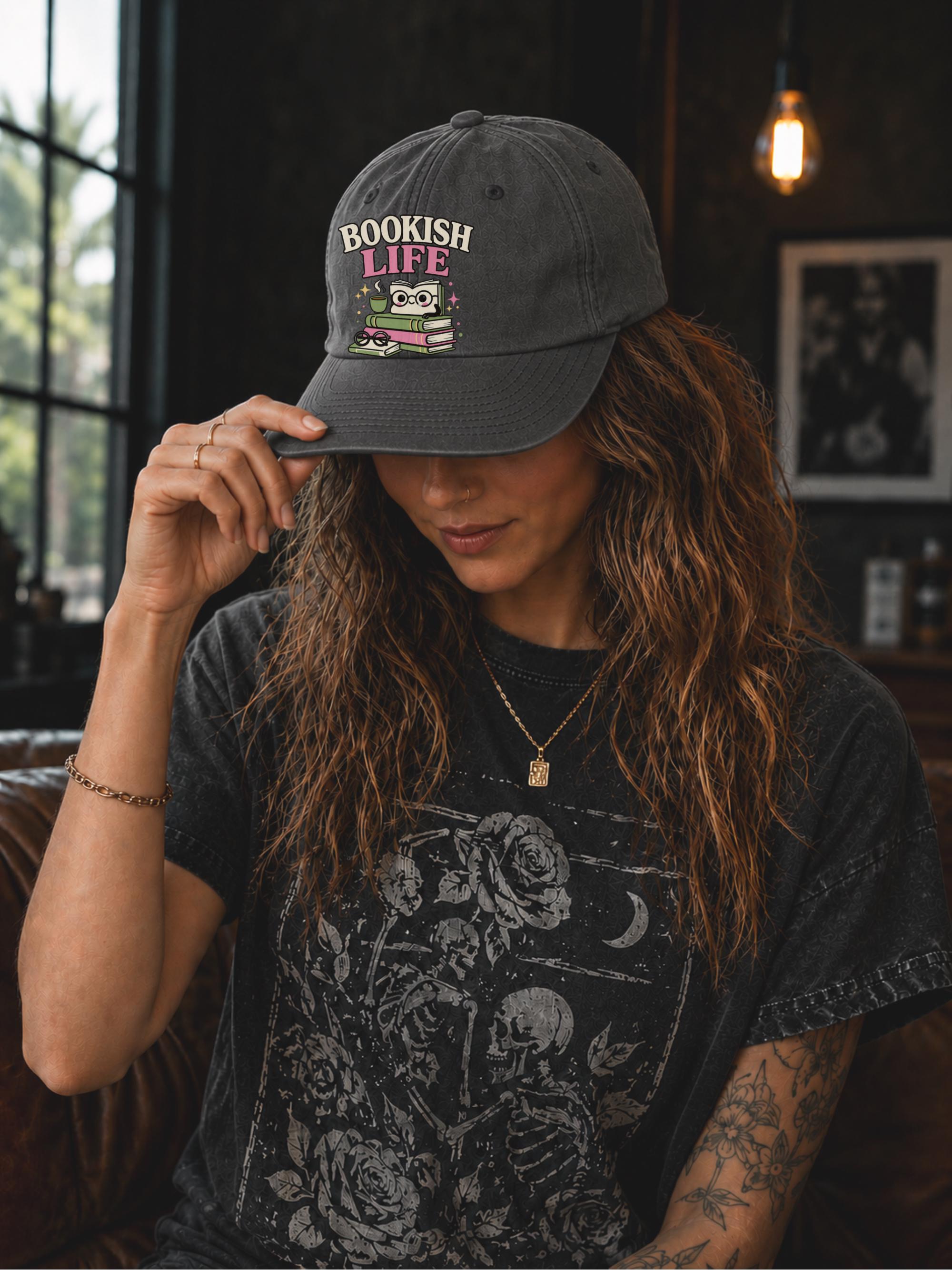 Woman wearing a dark grey classic dad cap featuring a colorful Bookish Life graphic with stacked books, glasses, and a playful reading-themed illustration.