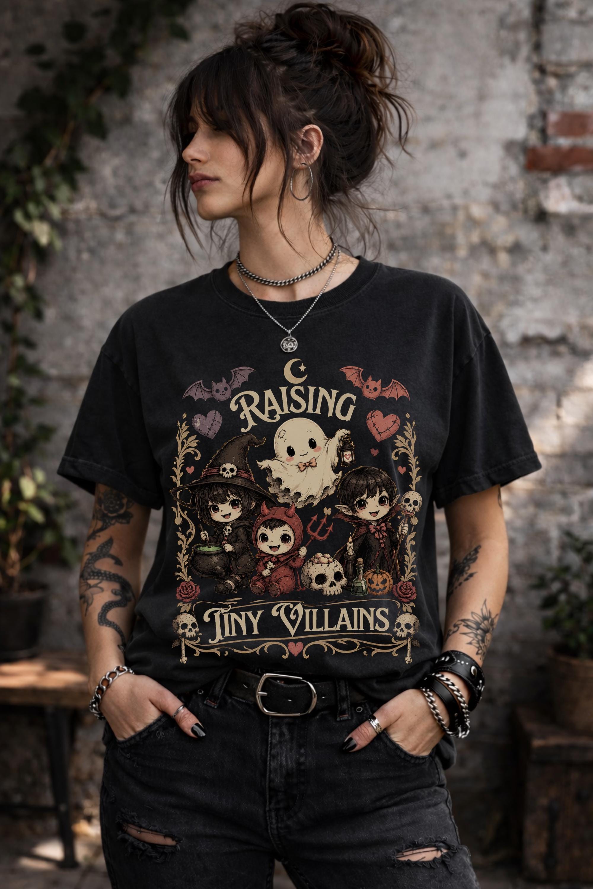 Raising Tiny Villains graphic t-shirt featuring cute ghost, witch, vampire, devil child, bats, skulls, hearts, and spooky gothic artwork.