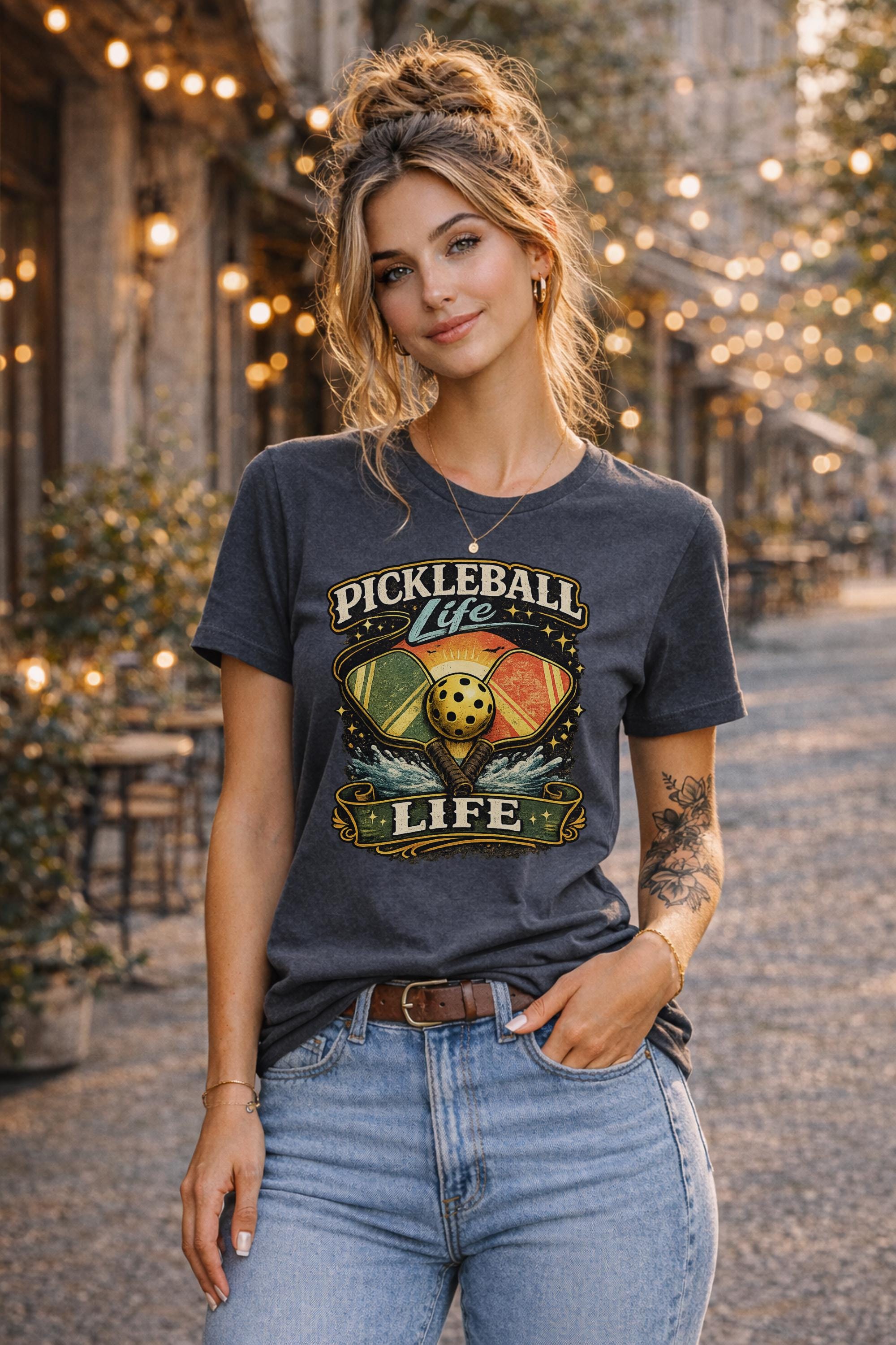 Woman wearing a Pickleball Life graphic t-shirt featuring colorful retro paddles, pickleball artwork, and vintage sports design in an outdoor street setting with string lights.