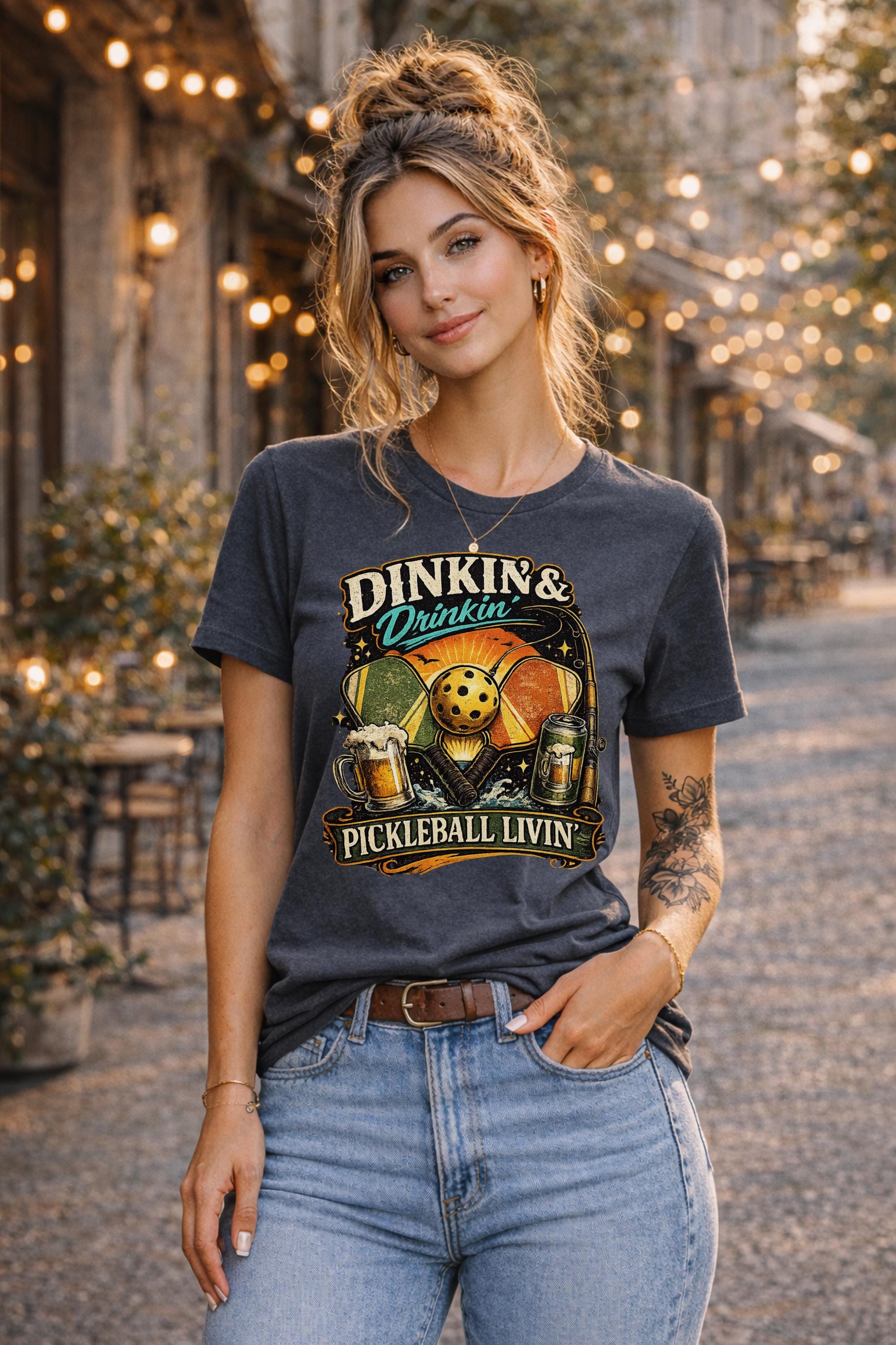Woman wearing a Dinkin & Drinkin Pickleball Livin graphic t-shirt featuring retro paddles, pickleball artwork, beer mugs, and vintage sunset design in an outdoor café setting with string lights.