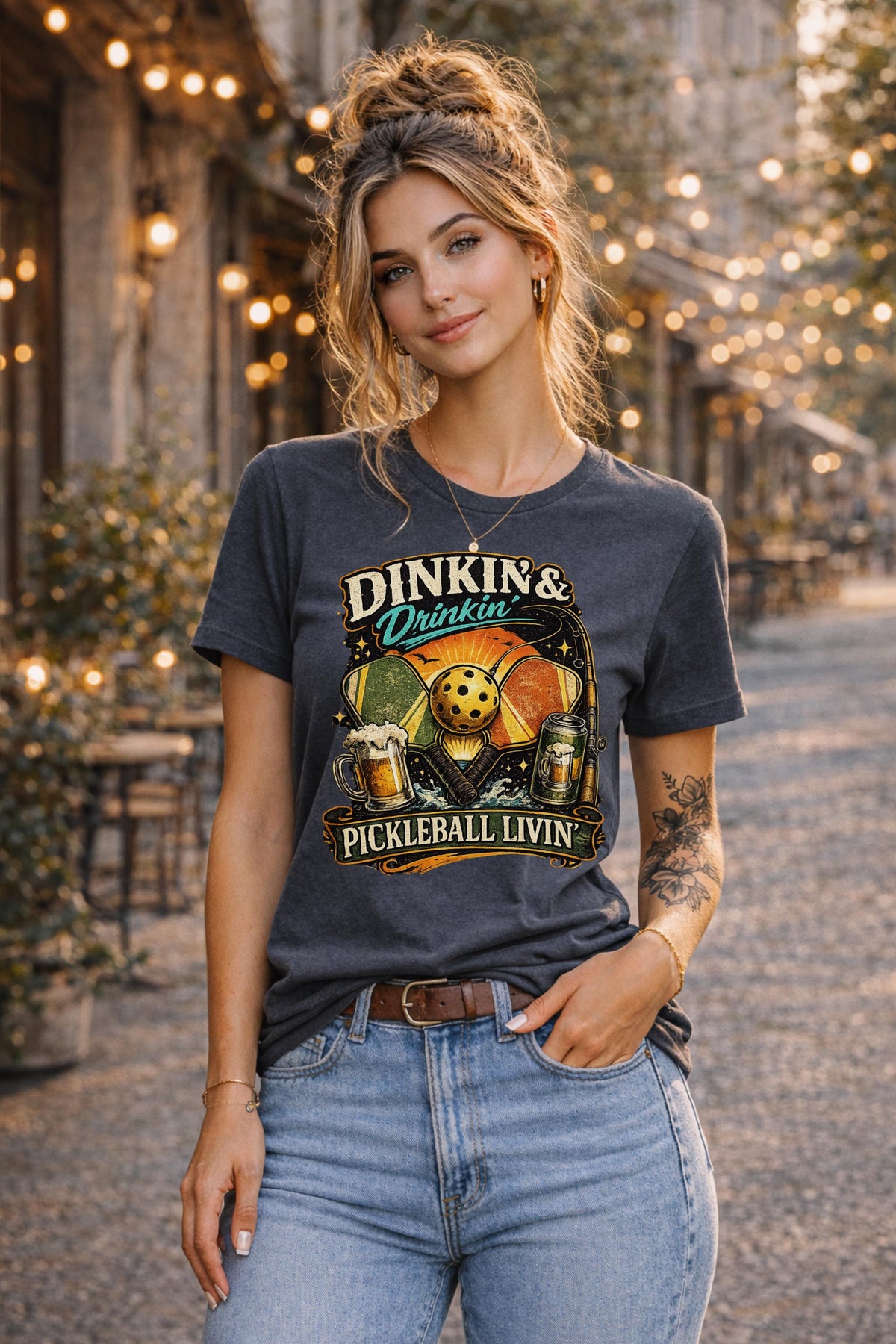 Woman wearing a Dinkin & Drinkin Pickleball Livin graphic t-shirt featuring retro paddles, pickleball artwork, beer mugs, and vintage sunset design in an outdoor café setting with string lights.