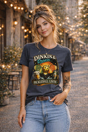 Woman wearing a Dinkin & Drinkin Pickleball Livin graphic t-shirt featuring retro paddles, pickleball artwork, beer mugs, and vintage sunset design in an outdoor café setting with string lights.