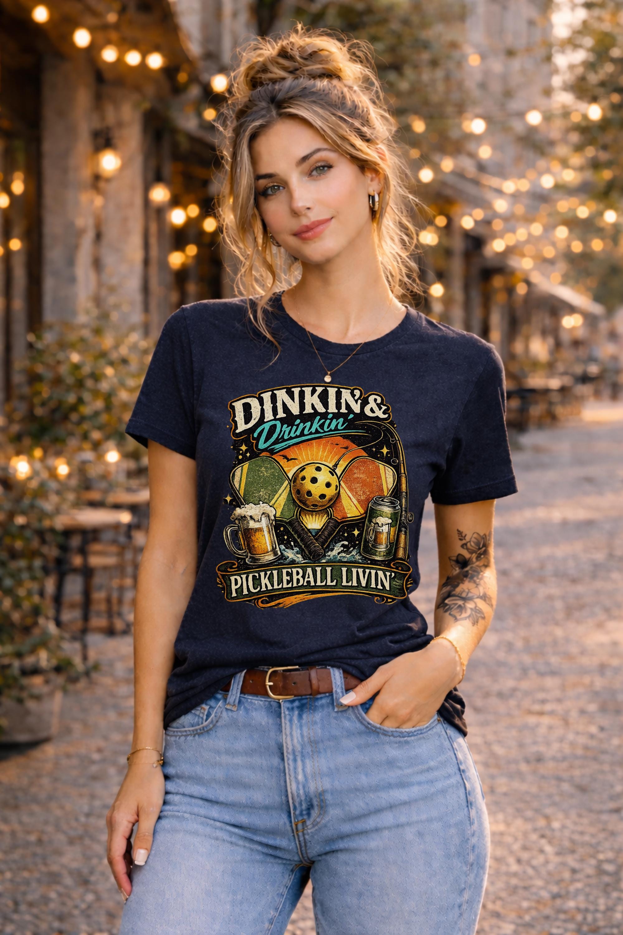 Woman wearing a Dinkin & Drinkin Pickleball Livin graphic t-shirt featuring retro paddles, pickleball artwork, beer mugs, and vintage sunset design in an outdoor café setting with string lights.