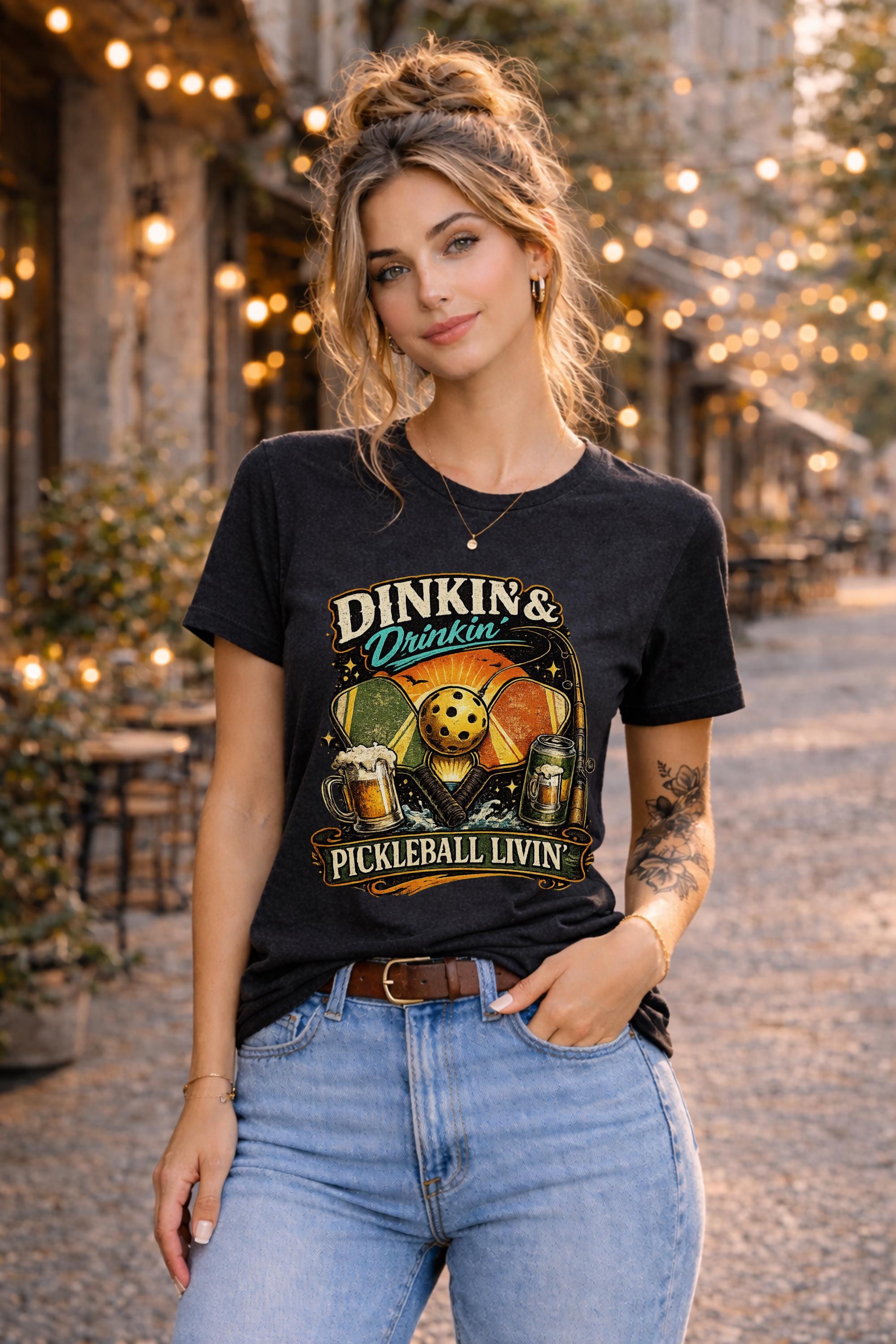 Woman wearing a Dinkin & Drinkin Pickleball Livin graphic t-shirt featuring retro paddles, pickleball artwork, beer mugs, and vintage sunset design in an outdoor café setting with string lights.