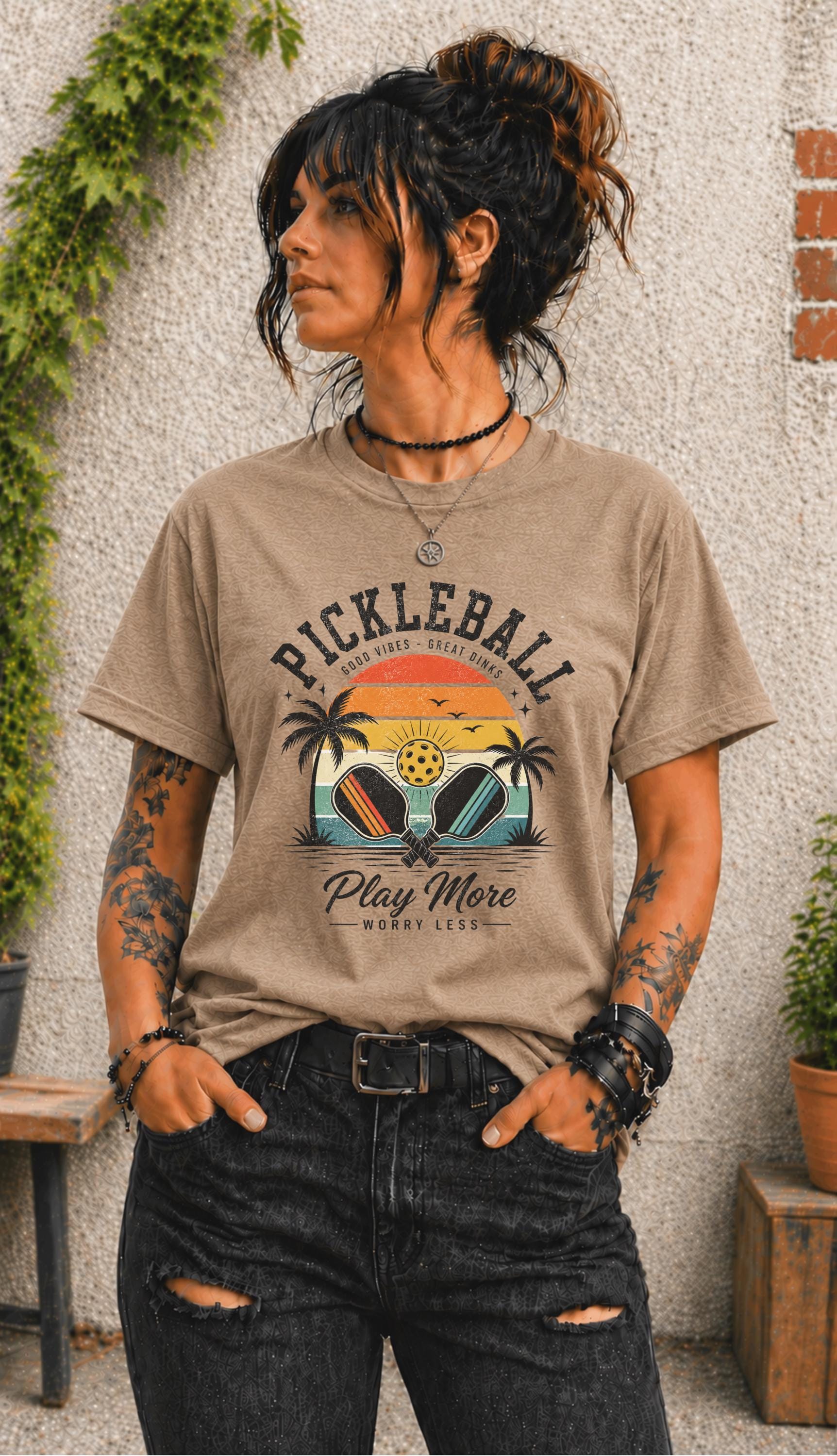 Woman wearing a Pickleball Play More Worry Less graphic t-shirt featuring retro paddles, tropical palm trees, vintage sunset artwork, and coastal-inspired pickleball design.