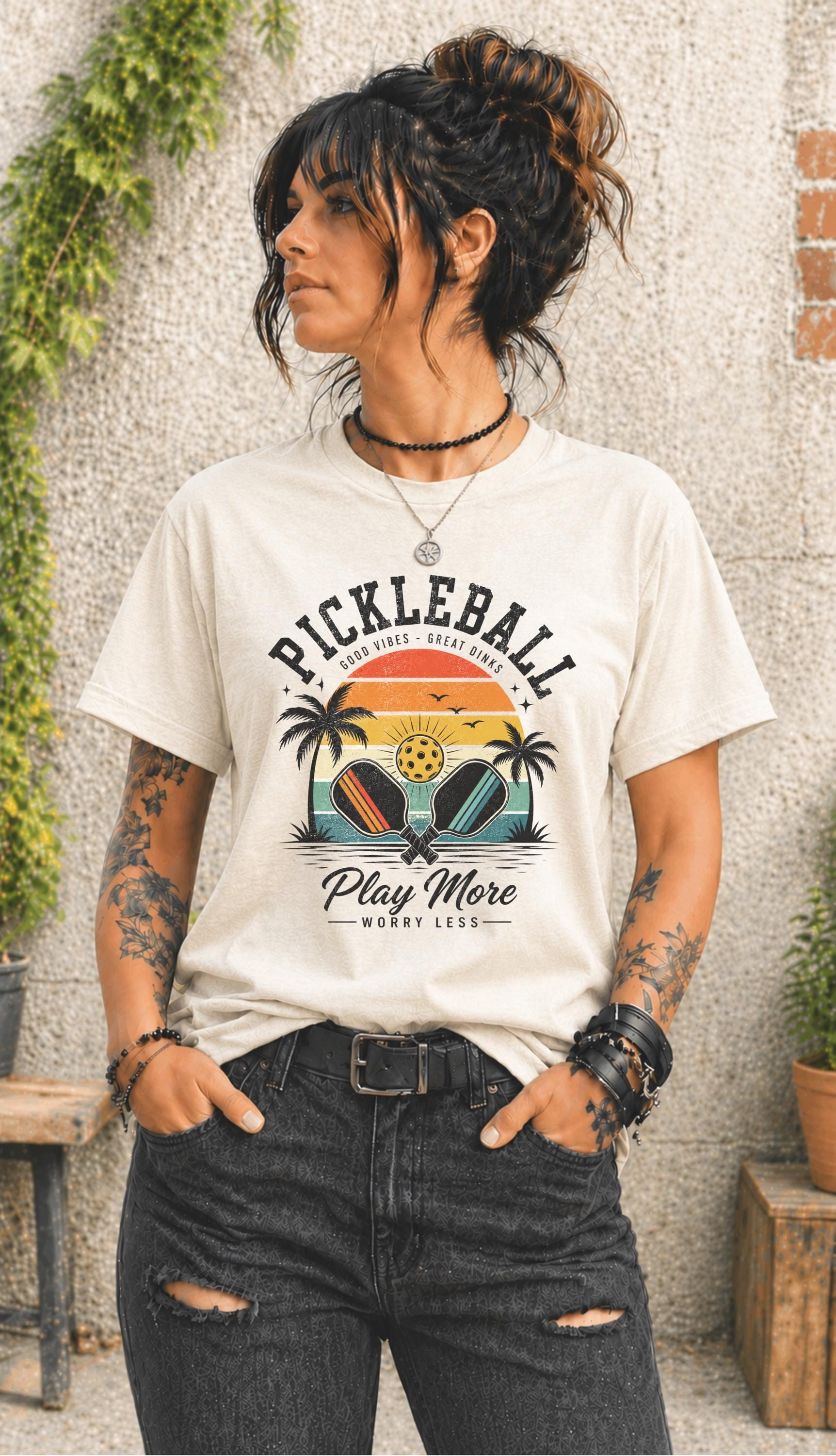 Woman wearing a Pickleball Play More Worry Less graphic t-shirt featuring retro paddles, tropical palm trees, vintage sunset artwork, and coastal-inspired pickleball design.