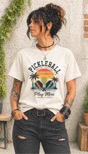 Woman wearing a Pickleball Play More Worry Less graphic t-shirt featuring retro paddles, tropical palm trees, vintage sunset artwork, and coastal-inspired pickleball design.