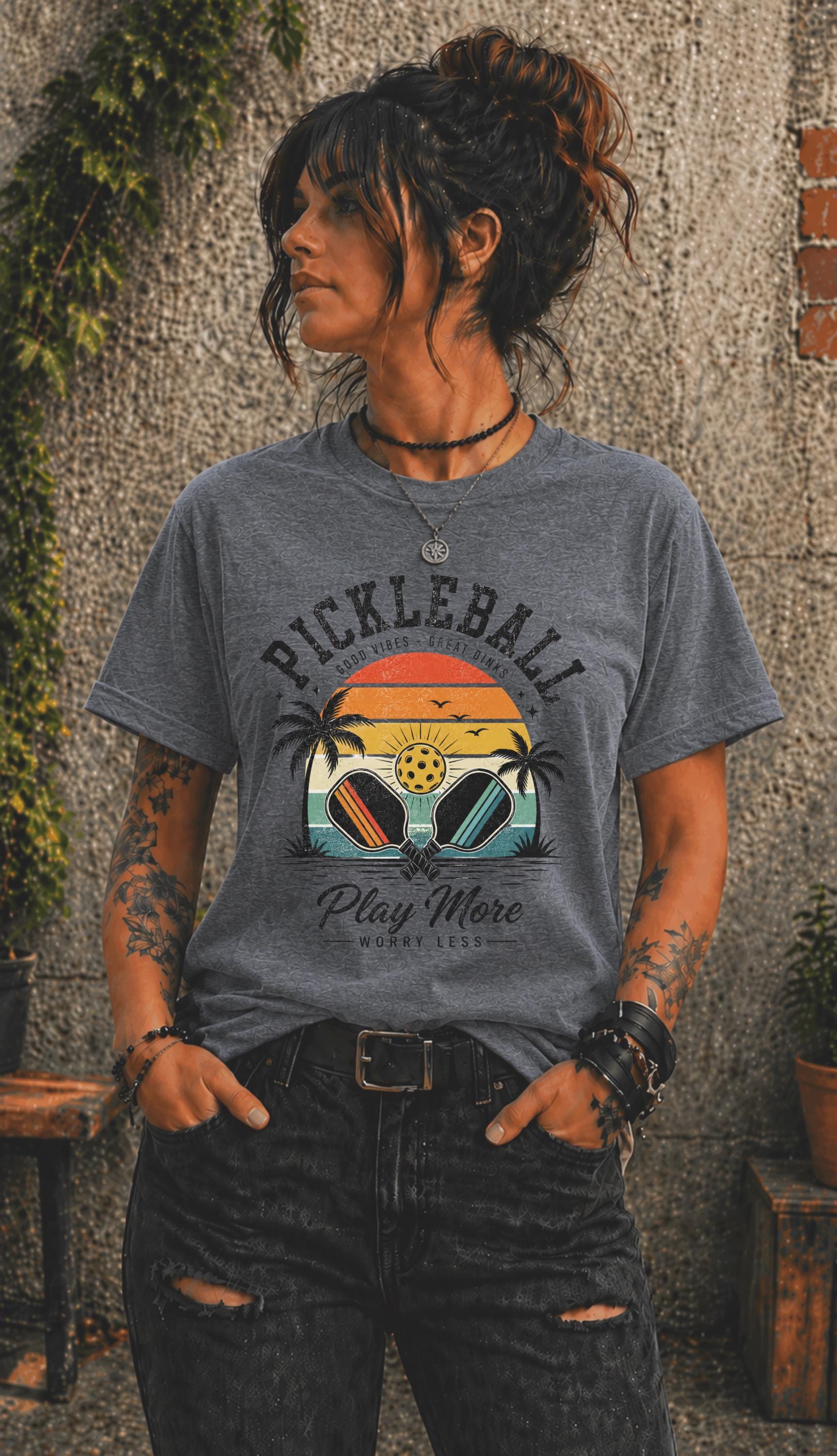 Woman wearing a Pickleball Play More Worry Less graphic t-shirt featuring retro paddles, tropical palm trees, vintage sunset artwork, and coastal-inspired pickleball design.