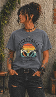 Woman wearing a Pickleball Play More Worry Less graphic t-shirt featuring retro paddles, tropical palm trees, vintage sunset artwork, and coastal-inspired pickleball design.