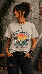 Woman wearing a Pickleball Play More Worry Less graphic t-shirt featuring retro paddles, tropical palm trees, vintage sunset artwork, and coastal-inspired pickleball design.