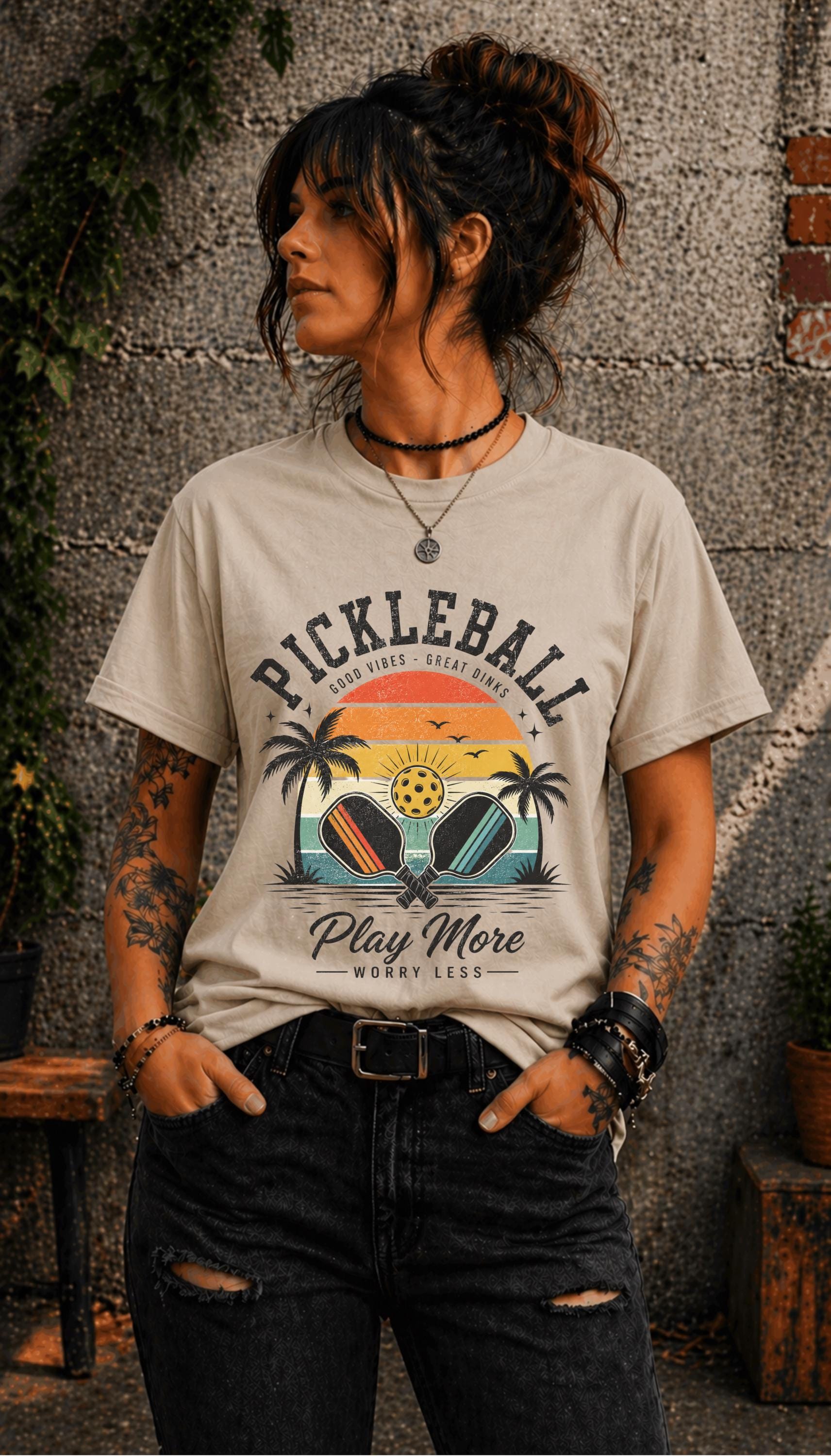 Woman wearing a Pickleball Play More Worry Less graphic t-shirt featuring retro paddles, tropical palm trees, vintage sunset artwork, and coastal-inspired pickleball design.