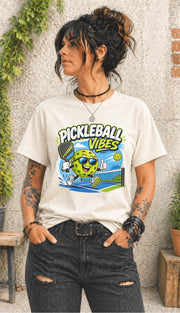 Woman wearing a Pickleball Vibes graphic t-shirt featuring a smiling pickleball character with sunglasses, paddle artwork, and colorful court-themed design.
