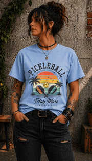 Woman wearing a light blue Pickleball Play More Worry Less graphic t-shirt featuring retro paddles, tropical palm trees, vintage sunset artwork, and coastal-inspired pickleball design.