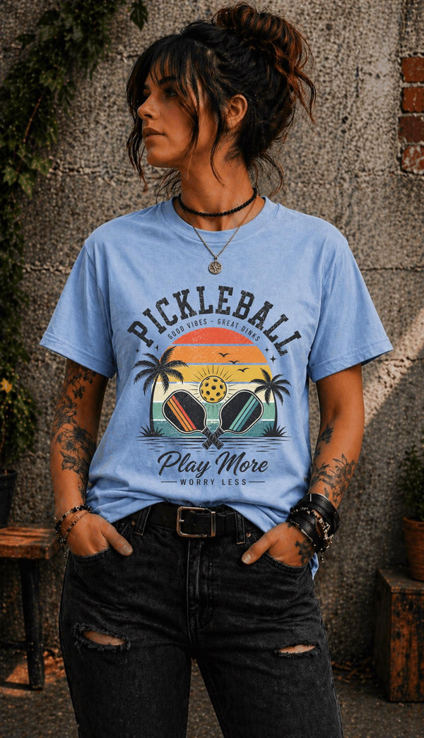 Woman wearing a light blue Pickleball Play More Worry Less graphic t-shirt featuring retro paddles, tropical palm trees, vintage sunset artwork, and coastal-inspired pickleball design.