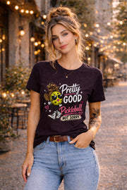 Woman wearing a pickleball graphic t-shirt featuring a stylish pickleball character with sunglasses, leopard-print bow, and the phrase "Pretty Good At Pickleball Not Sorry" in pink and white lettering.