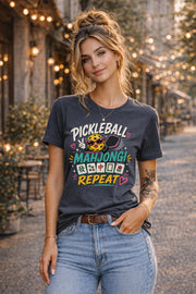 Woman wearing a graphic t-shirt featuring a colorful "Pickleball Mahjong Repeat" design with pickleball character, mahjong tiles, hearts, and retro-inspired artwork.
