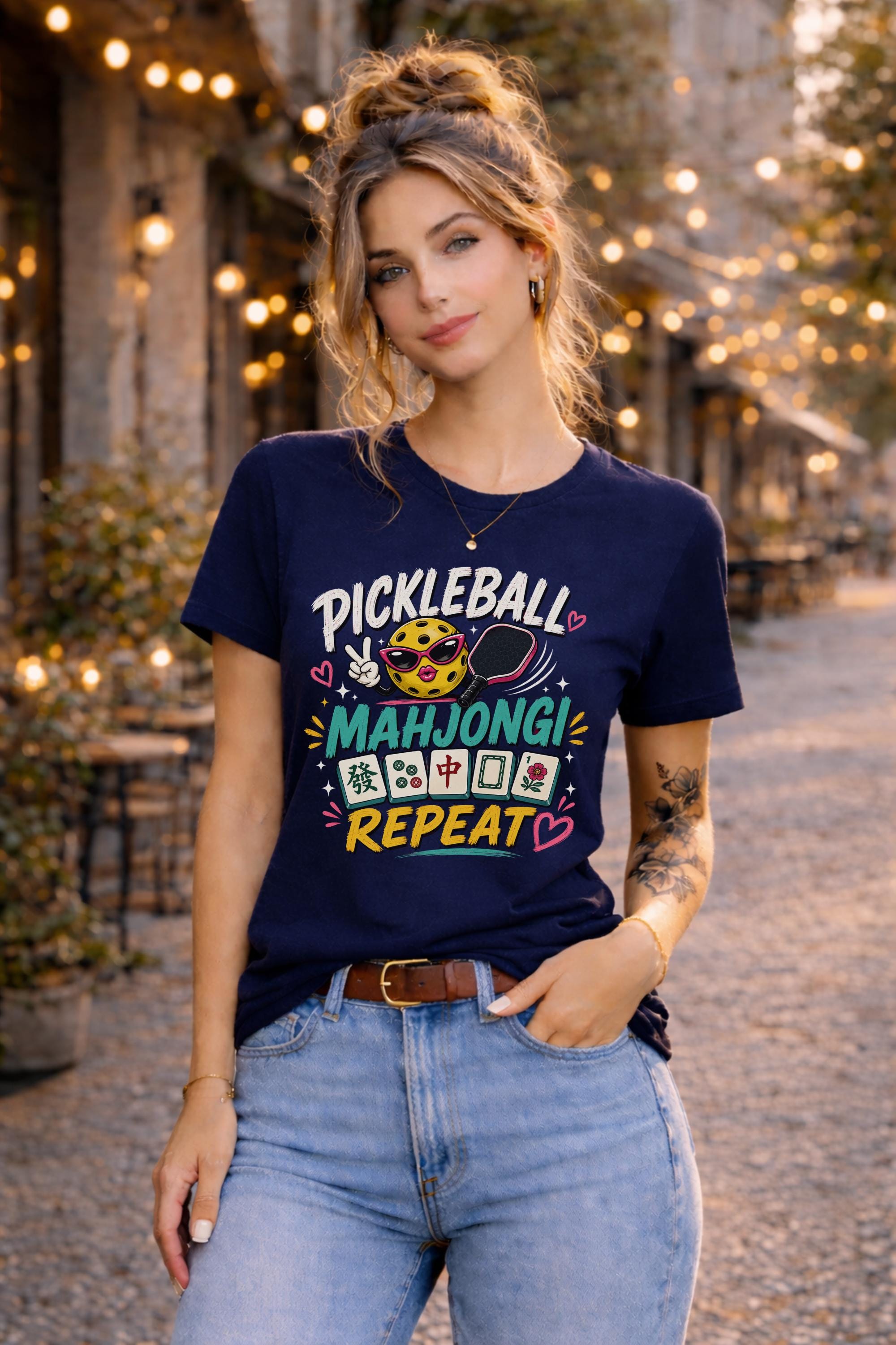 Woman wearing a graphic t-shirt featuring a colorful "Pickleball Mahjong Repeat" design with pickleball character, mahjong tiles, hearts, and retro-inspired artwork.