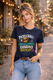 Woman wearing a graphic t-shirt featuring a colorful "Pickleball Mahjong Repeat" design with pickleball character, mahjong tiles, hearts, and retro-inspired artwork.