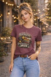 Woman wearing a graphic t-shirt featuring a Living That Bookish Pickleball Life design with books, coffee cup, pickleball paddles, floral accents, and vintage-inspired artwork.