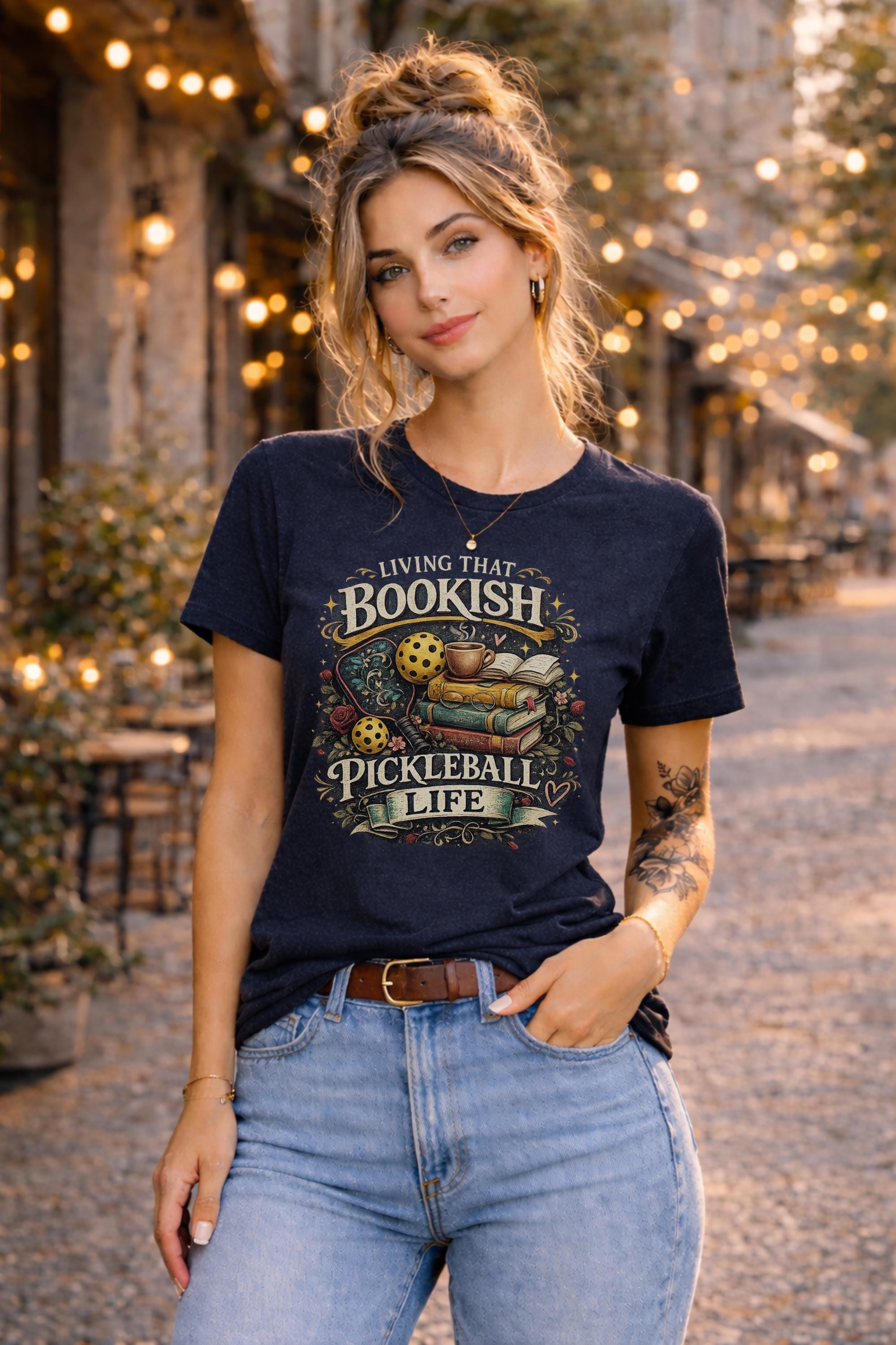 Woman wearing a graphic t-shirt featuring a Living That Bookish Pickleball Life design with books, coffee cup, pickleball paddles, floral accents, and vintage-inspired artwork.
