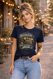 Woman wearing a graphic t-shirt featuring a Living That Bookish Pickleball Life design with books, coffee cup, pickleball paddles, floral accents, and vintage-inspired artwork.