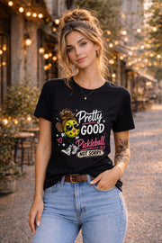 Woman wearing a black pickleball graphic t-shirt featuring a stylish pickleball character with sunglasses, leopard-print bow, and the phrase "Pretty Good At Pickleball Not Sorry" in pink and white lettering.