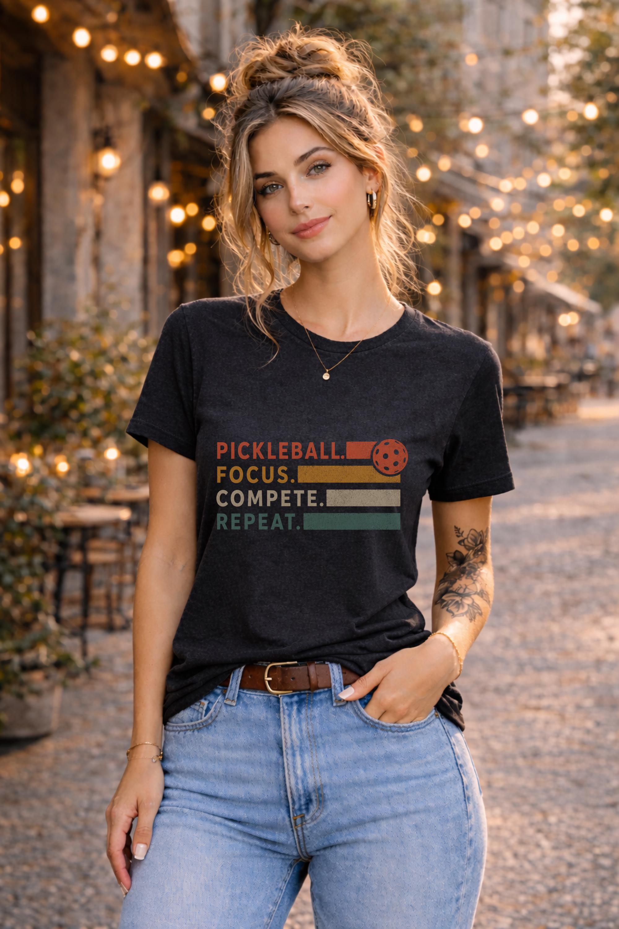 Woman wearing a graphic t-shirt featuring a vintage Pickleball Focus Compete Repeat design with retro stripes, pickleball icon, and motivational athletic typography.