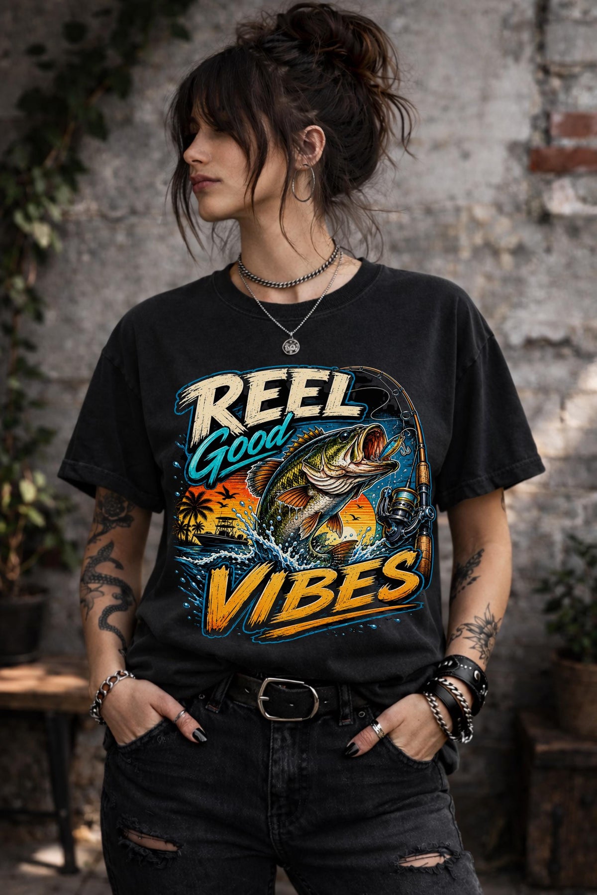 Woman wearing a graphic t-shirt featuring a jumping bass fish, fishing rod, tropical sunset scene, and Reel Good Vibes lettering.