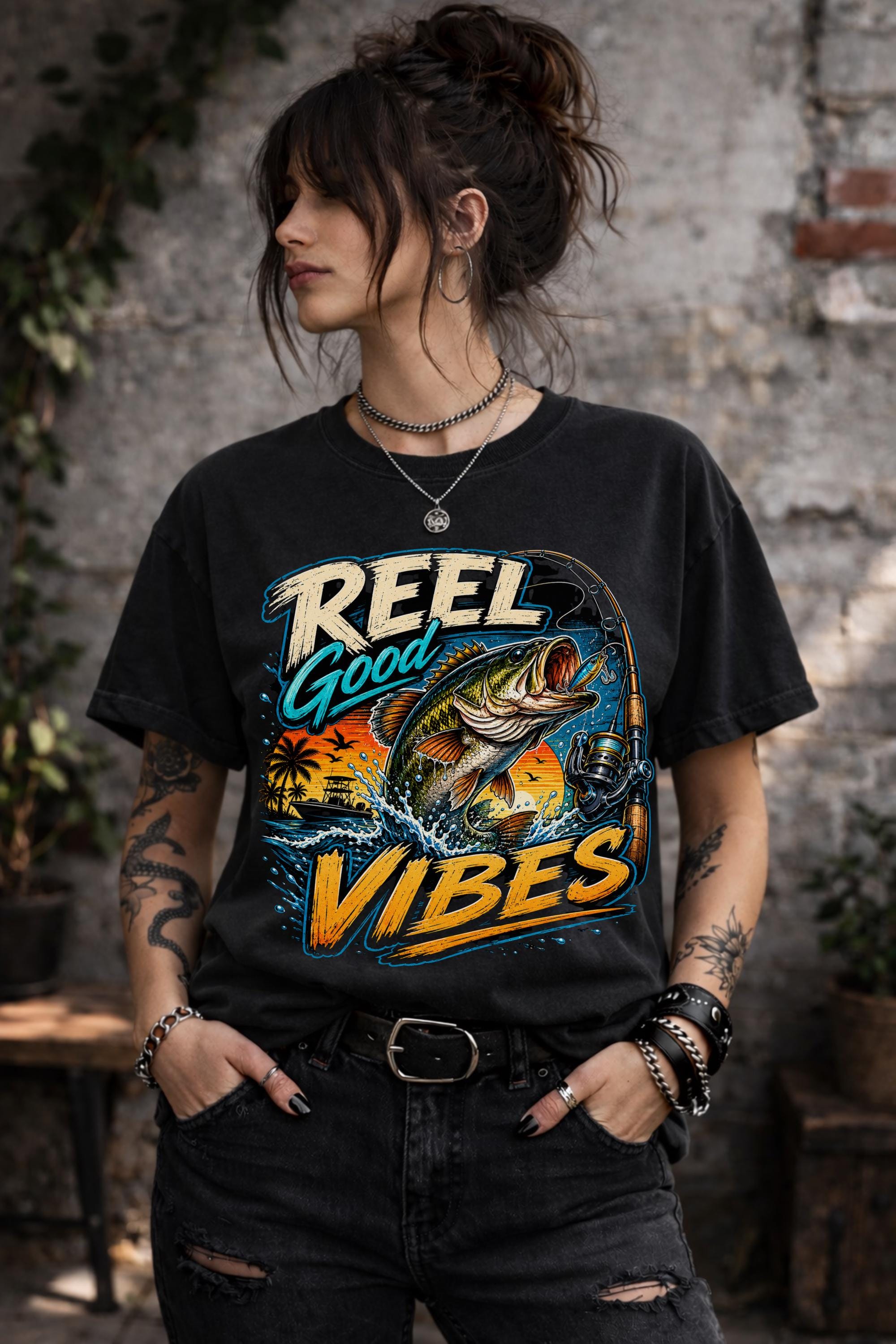 Woman wearing a graphic t-shirt featuring a jumping bass fish, fishing rod, tropical sunset scene, and Reel Good Vibes lettering.