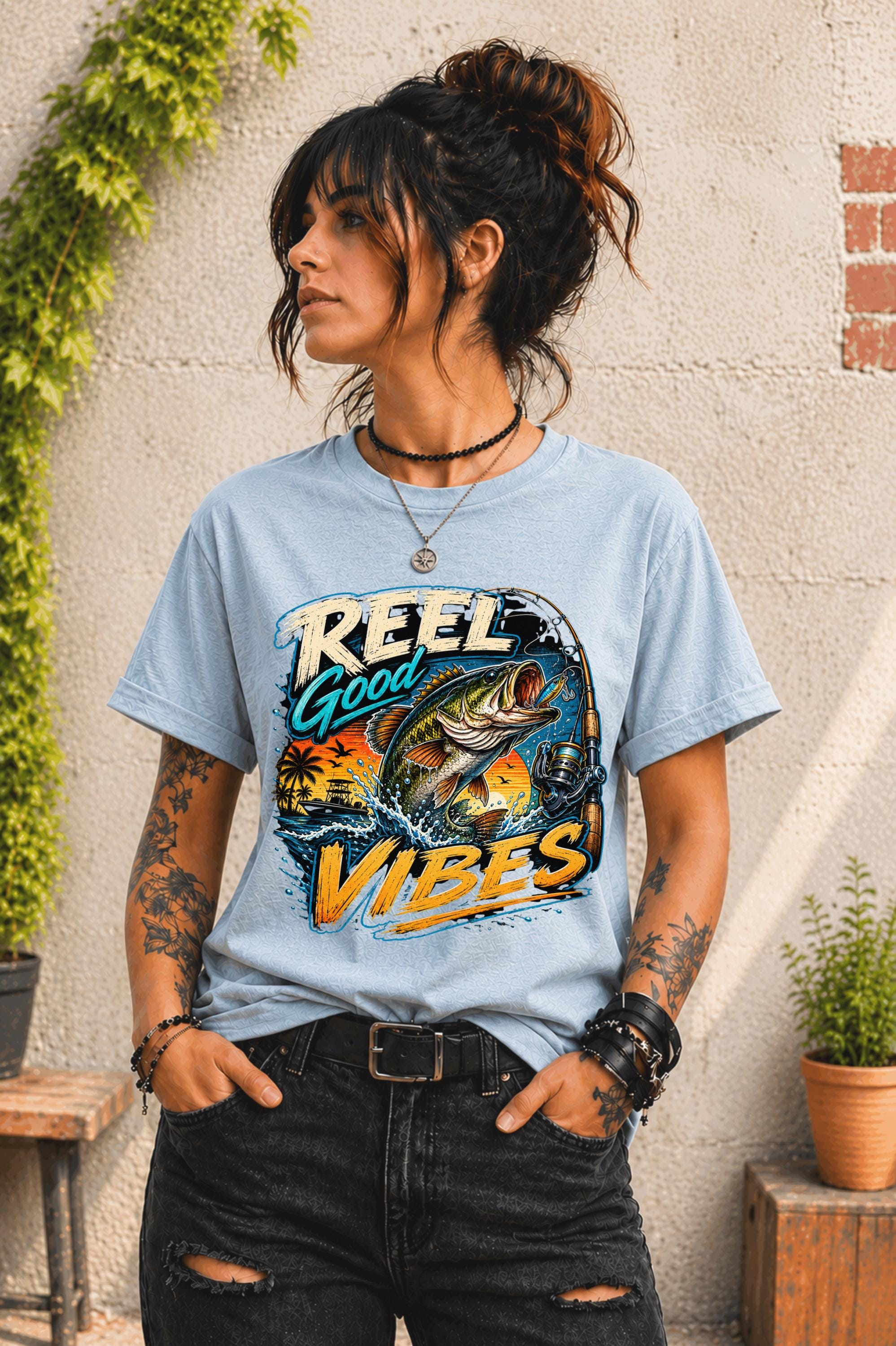 Woman wearing a graphic t-shirt featuring a jumping bass fish, fishing rod, tropical sunset scene, and Reel Good Vibes lettering.