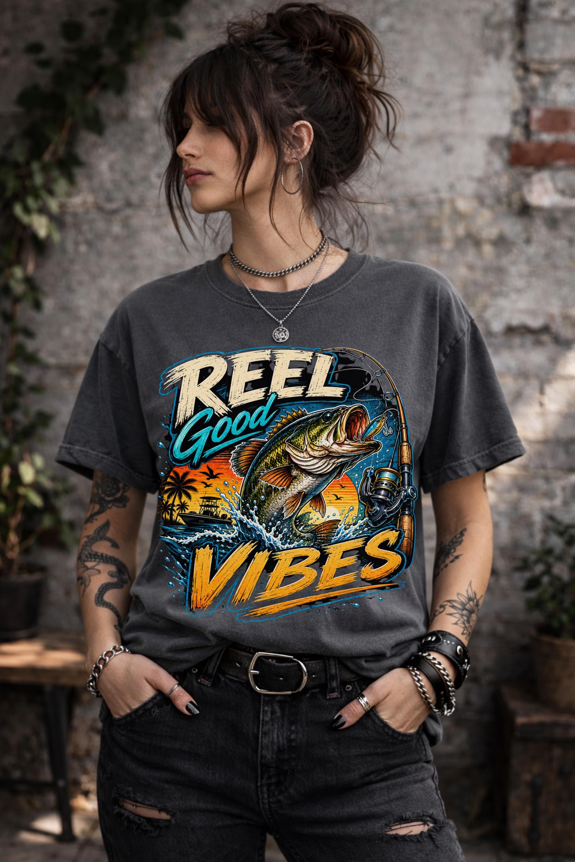 Woman wearing a graphic t-shirt featuring a jumping bass fish, fishing rod, tropical sunset scene, and Reel Good Vibes lettering.