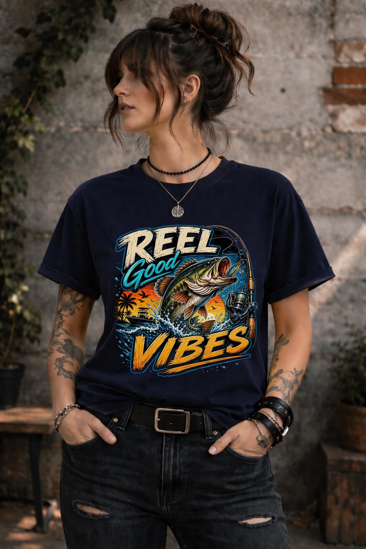 Woman wearing a graphic t-shirt featuring a jumping bass fish, fishing rod, tropical sunset scene, and Reel Good Vibes lettering.