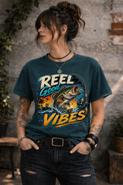 Woman wearing a graphic t-shirt featuring a jumping bass fish, fishing rod, tropical sunset scene, and Reel Good Vibes lettering.