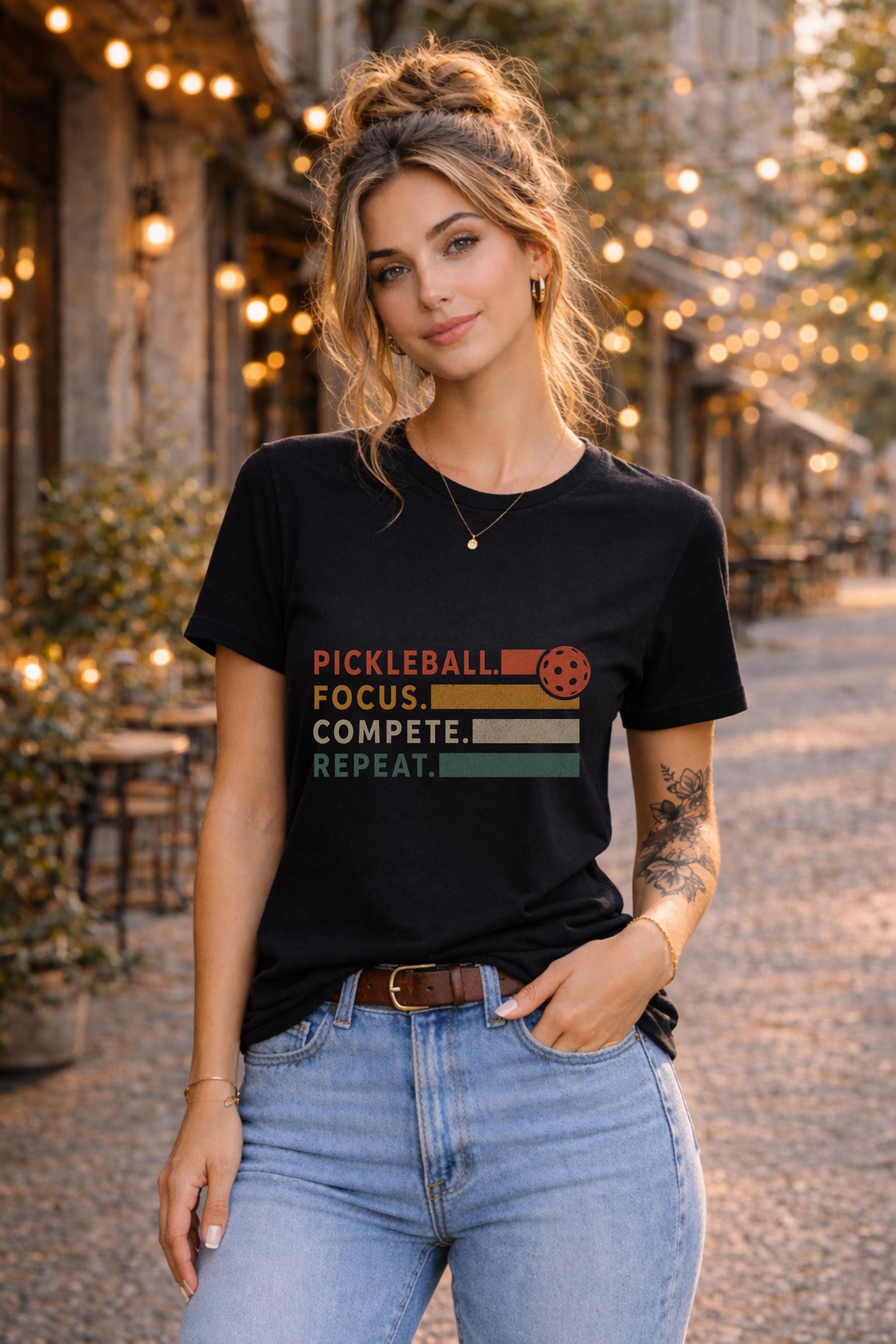 Woman wearing a black graphic t-shirt featuring a vintage Pickleball Focus Compete Repeat design with retro stripes, pickleball icon, and motivational athletic typography.