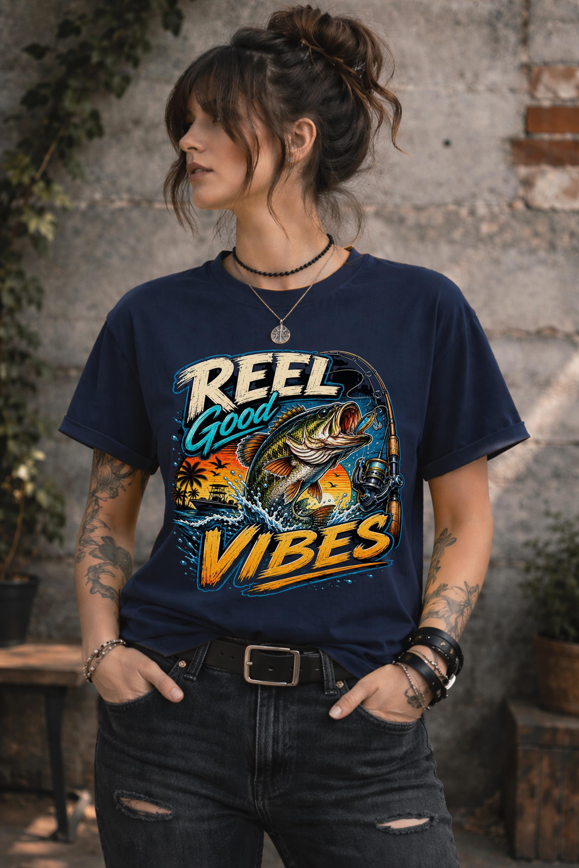 Woman wearing a navy graphic t-shirt featuring a jumping bass fish, fishing rod, tropical sunset scene, and Reel Good Vibes lettering.