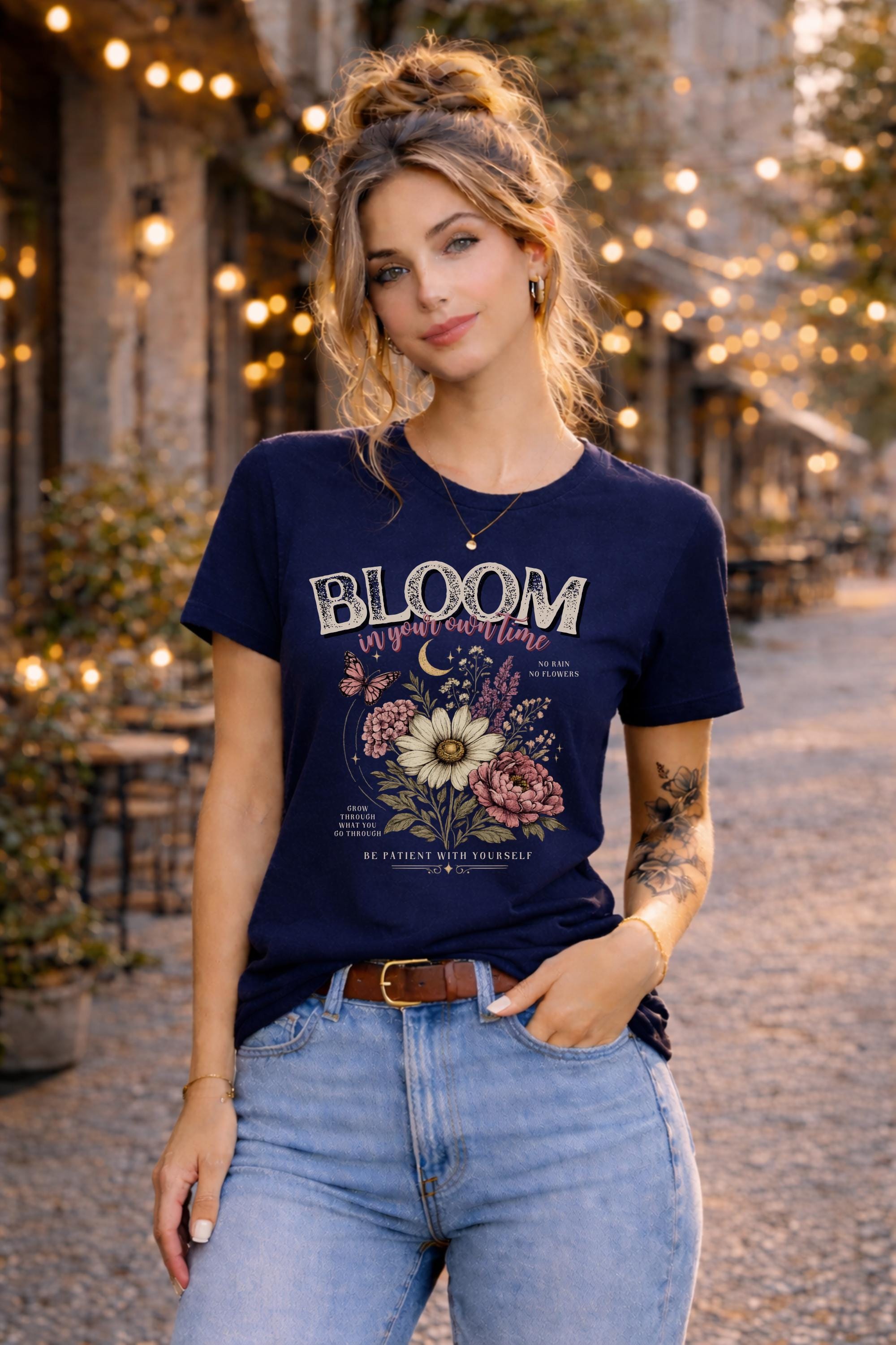 Woman wearing a floral inspirational t-shirt featuring the phrase "Bloom In Your Own Time" with vintage flowers, butterflies, moon accents, and self-love encouragement messages.