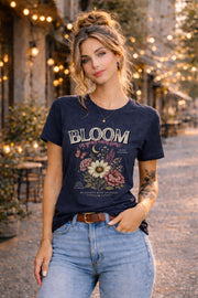 Woman wearing a floral inspirational t-shirt featuring the phrase "Bloom In Your Own Time" with vintage flowers, butterflies, moon accents, and self-love encouragement messages.