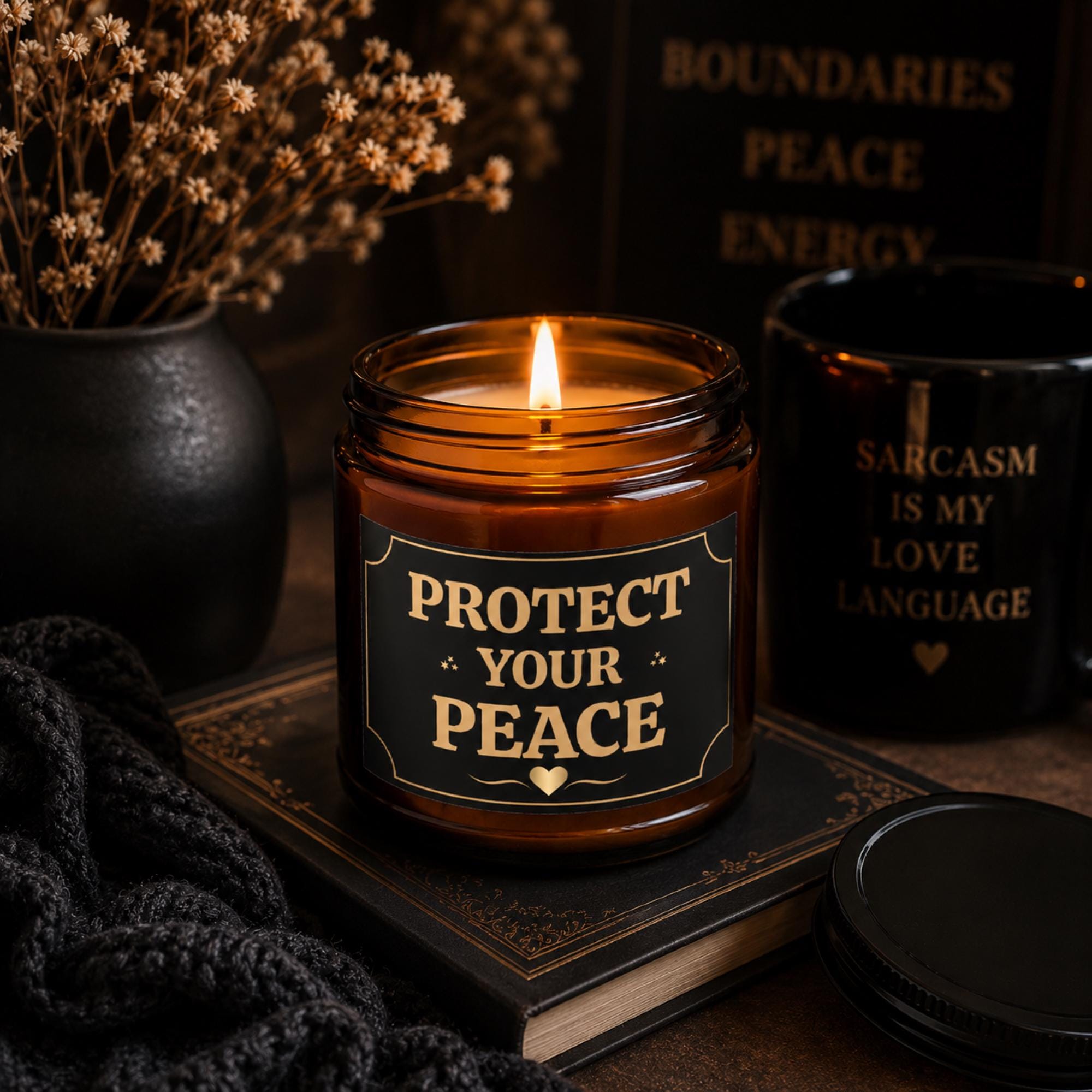 Amber glass candle with black and gold Protect Your Peace label in moody dark aesthetic setting with books and dried florals.
