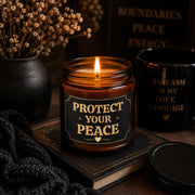 Amber glass candle with black and gold Protect Your Peace label in moody dark aesthetic setting with books and dried florals.