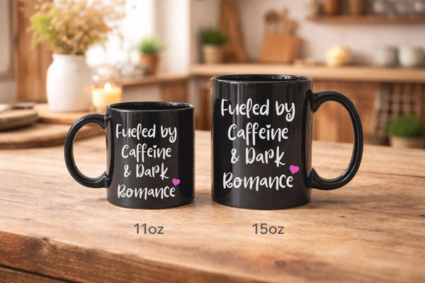 11oz and 15oz black ceramic mugs featuring the phrase "Fueled by Caffeine & Dark Romance" displayed on a rustic wooden table in a cozy kitchen setting.