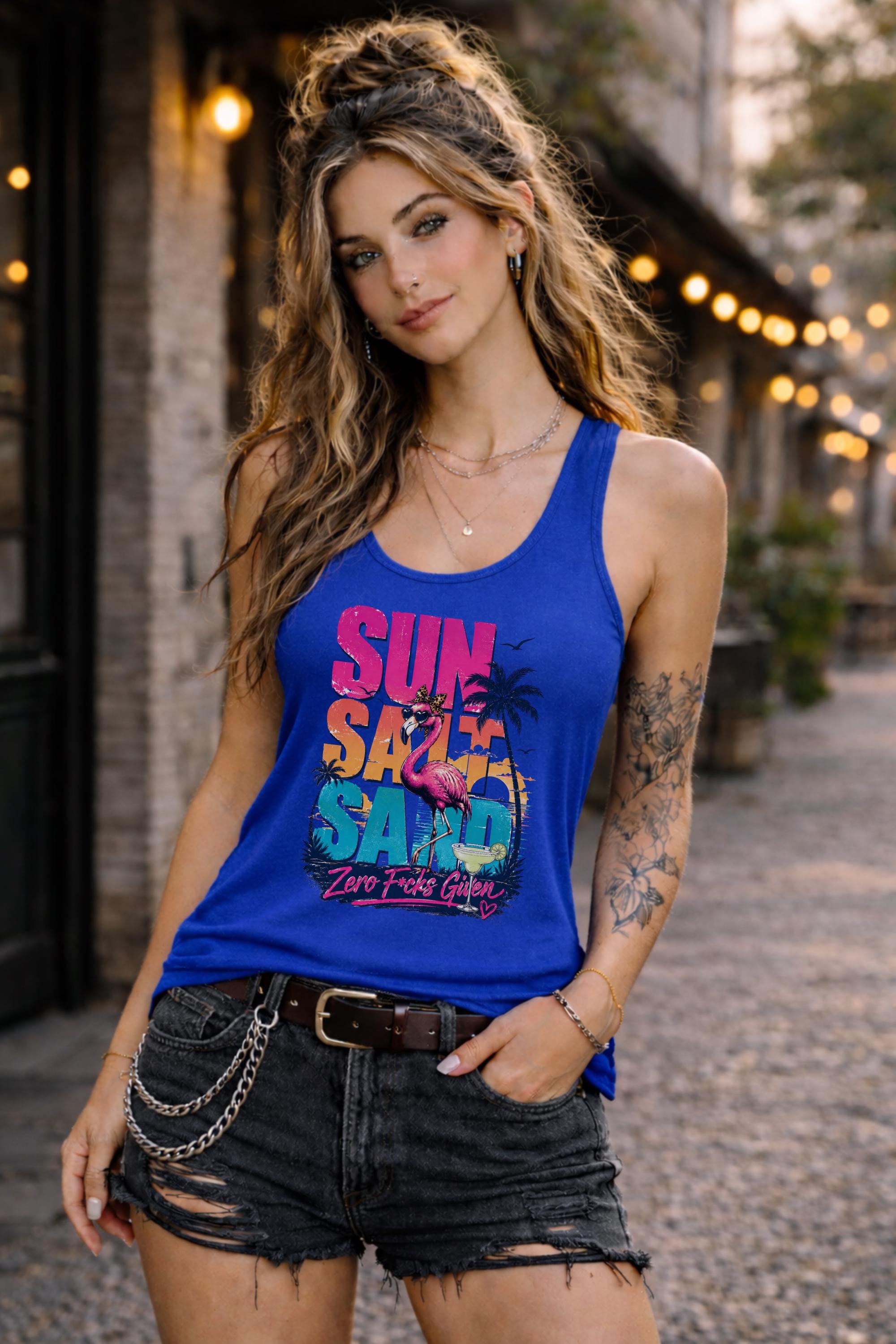 Woman modeling dark tank top featuring colorful retro flamingo graphic with Sun Sand Salad Zero Fcks Given summer vacation design against urban industrial background.