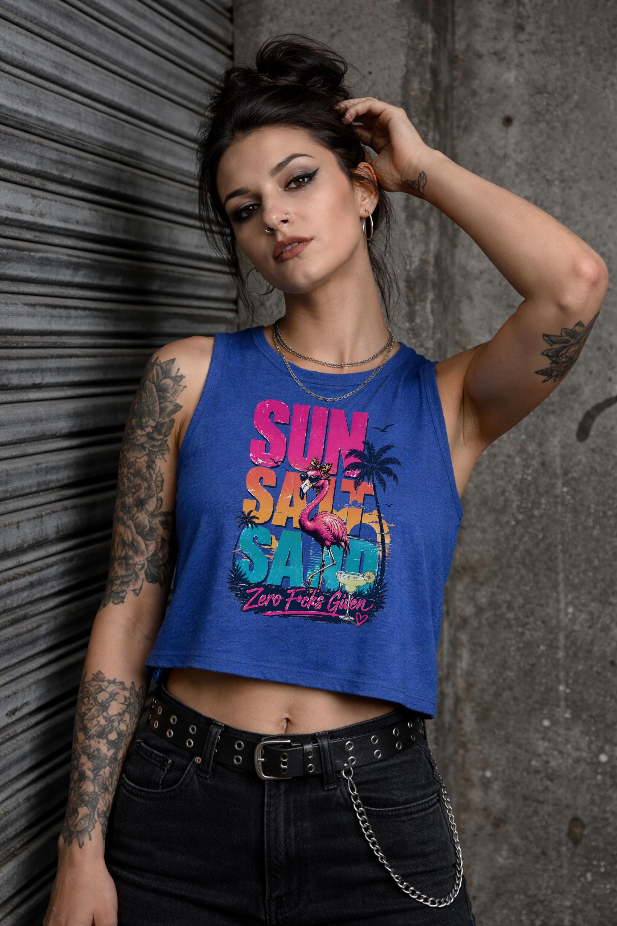 Woman modeling crop tank top featuring colorful retro flamingo graphic with Sun Sand Salad Zero Fcks Given summer vacation design against urban industrial background.