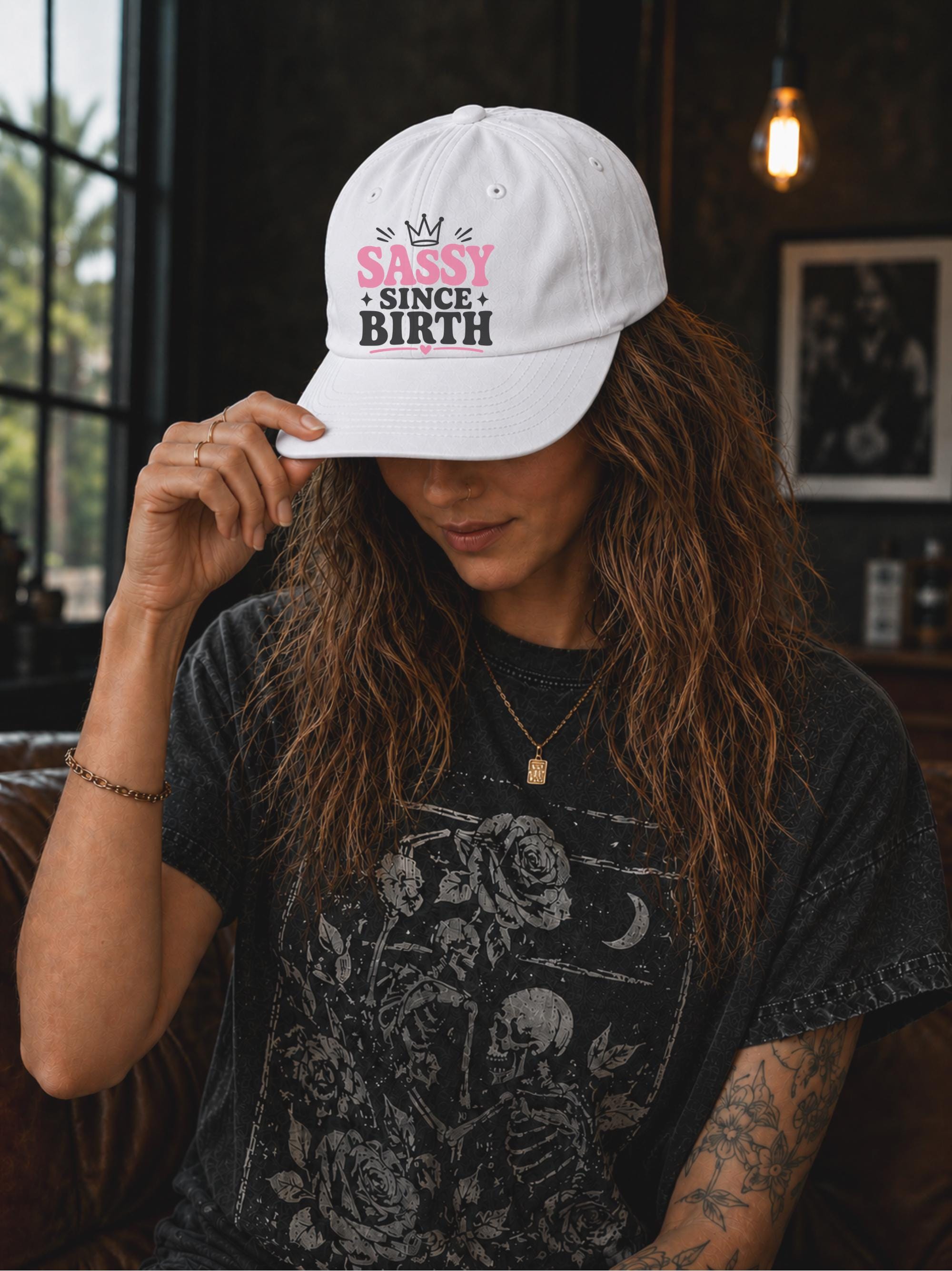 Classic dad cap featuring a pink and white "Sassy Since Birth" graphic with crown artwork, styled in a casual fashion setting.