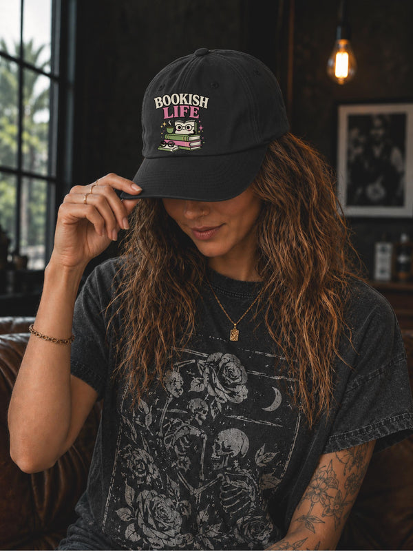 Woman wearing a black classic dad cap featuring a colorful Bookish Life graphic with stacked books, glasses, and a playful reading-themed illustration.