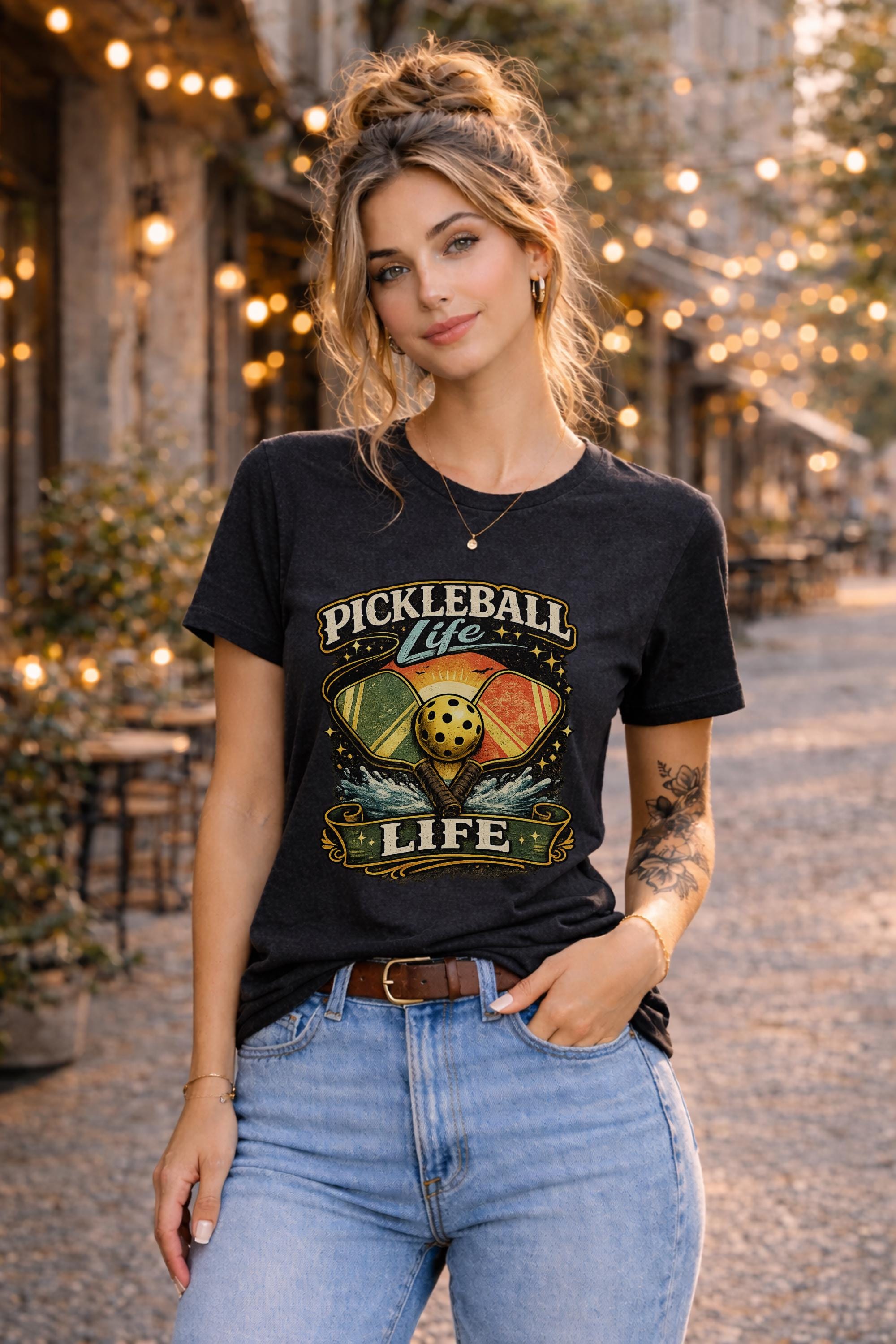 Woman wearing a Pickleball Life graphic t-shirt featuring colorful retro paddles, pickleball artwork, and vintage sports design in an outdoor street setting with string lights.