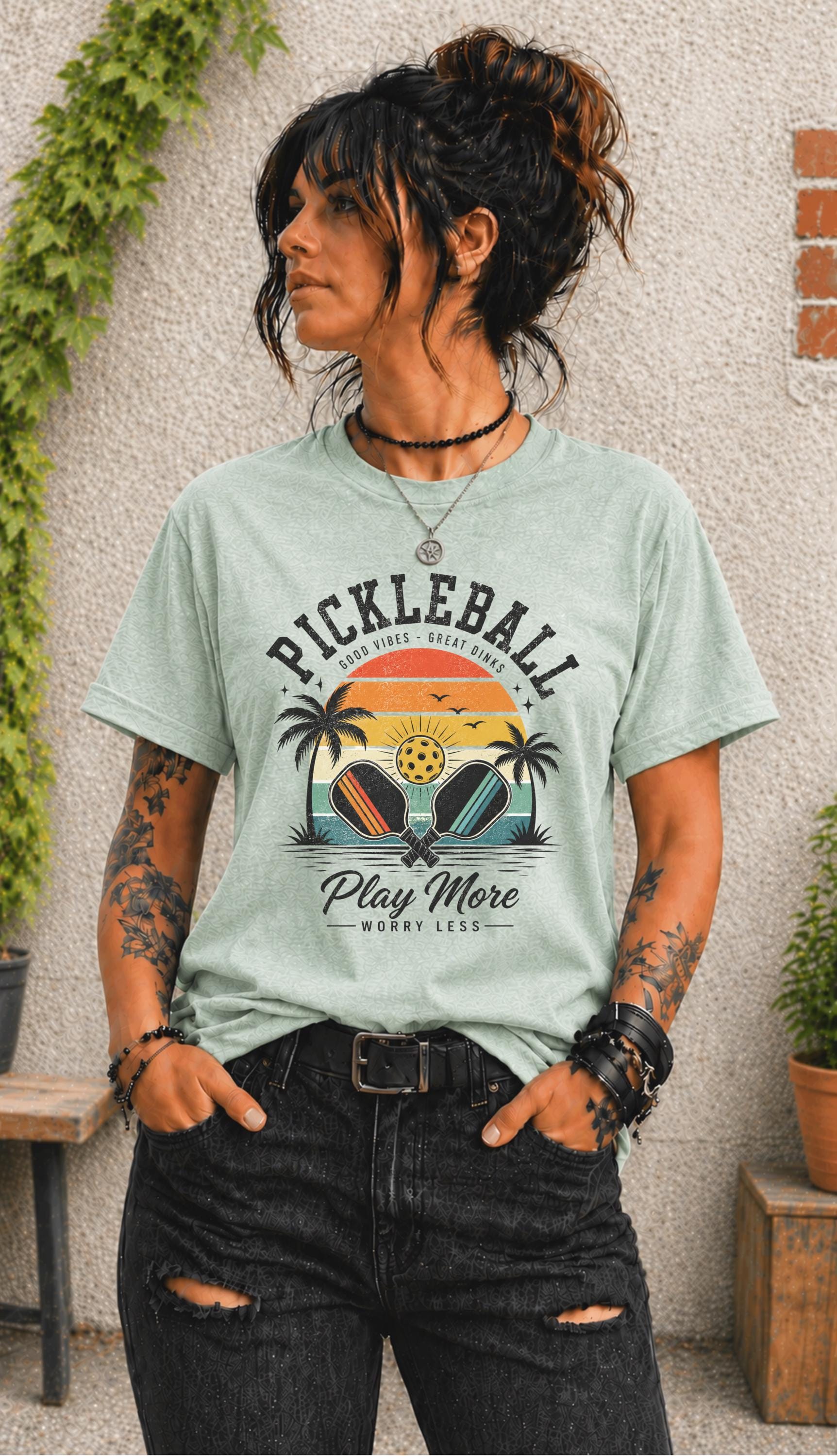 Woman wearing a Pickleball Play More Worry Less graphic t-shirt featuring retro paddles, tropical palm trees, vintage sunset artwork, and coastal-inspired pickleball design.