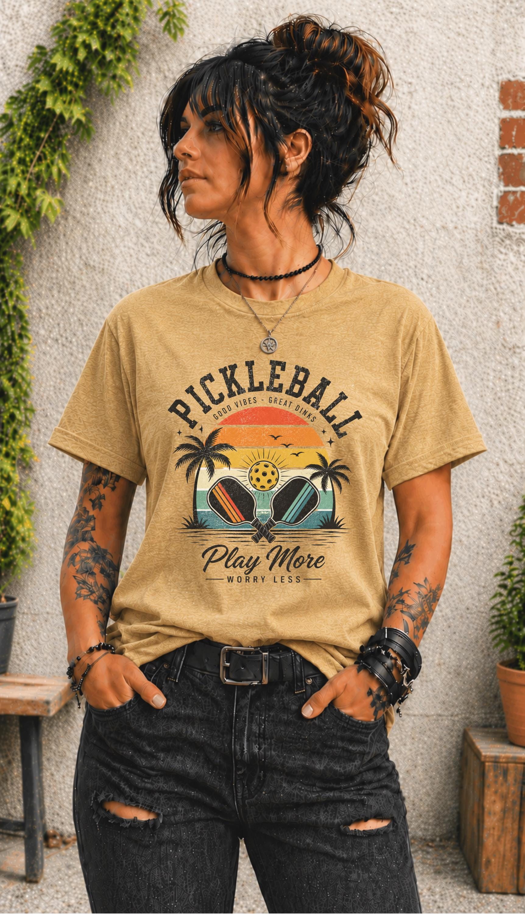 Woman wearing a Pickleball Play More Worry Less graphic t-shirt featuring retro paddles, tropical palm trees, vintage sunset artwork, and coastal-inspired pickleball design.