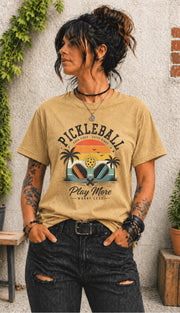 Woman wearing a Pickleball Play More Worry Less graphic t-shirt featuring retro paddles, tropical palm trees, vintage sunset artwork, and coastal-inspired pickleball design.