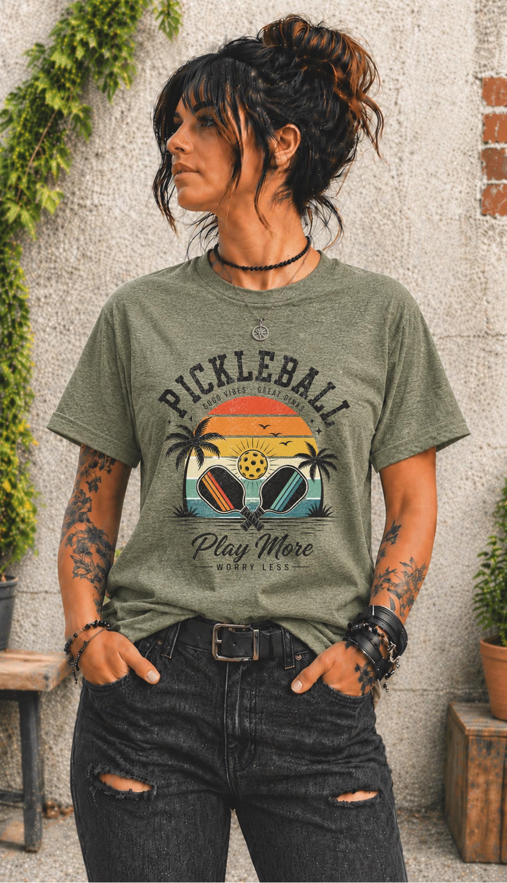 Woman wearing a Pickleball Play More Worry Less graphic t-shirt featuring retro paddles, tropical palm trees, vintage sunset artwork, and coastal-inspired pickleball design.
